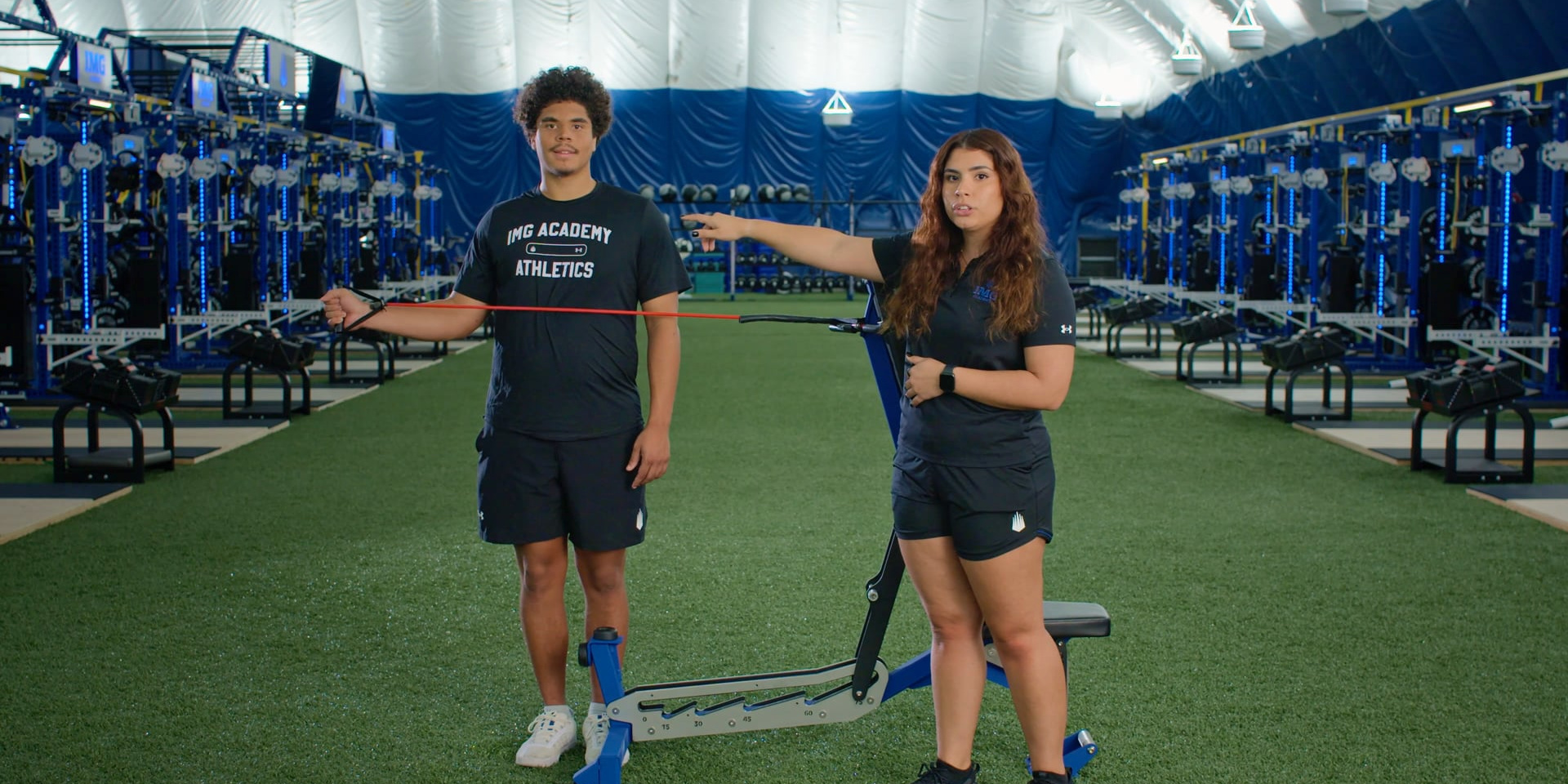 A trainer guides an athlete through a resistance-band shoulder exercise.