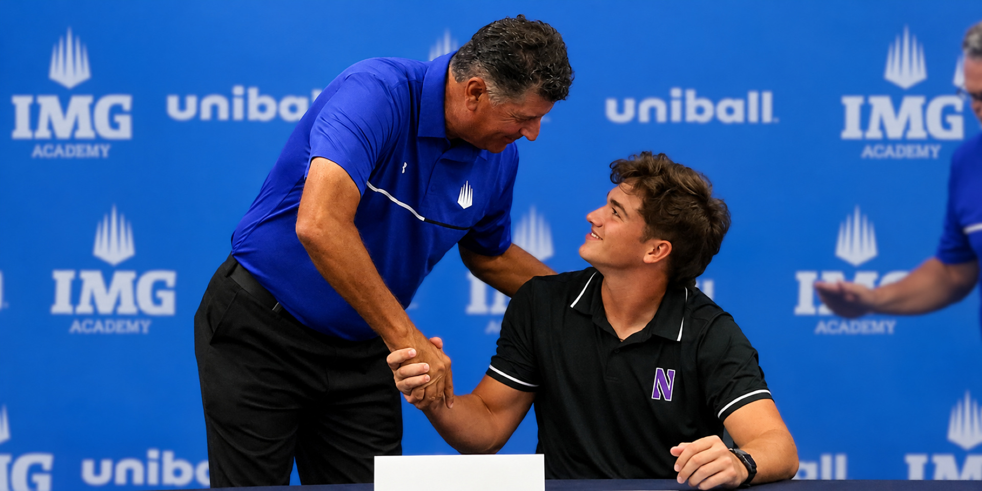 A seated student-athlete shakes hands with an adult at a signing table in front of an IMG Academy backdrop.