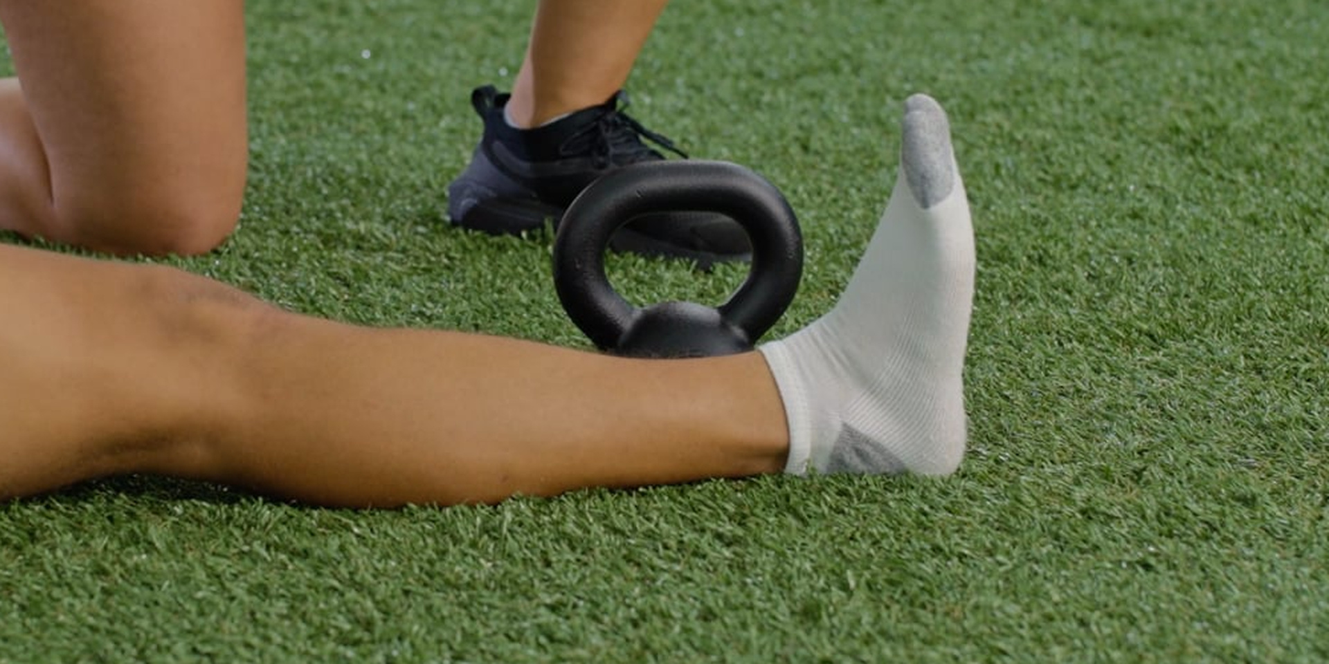 A close view shows an athlete lifting an extended leg over a kettlebell on turf.