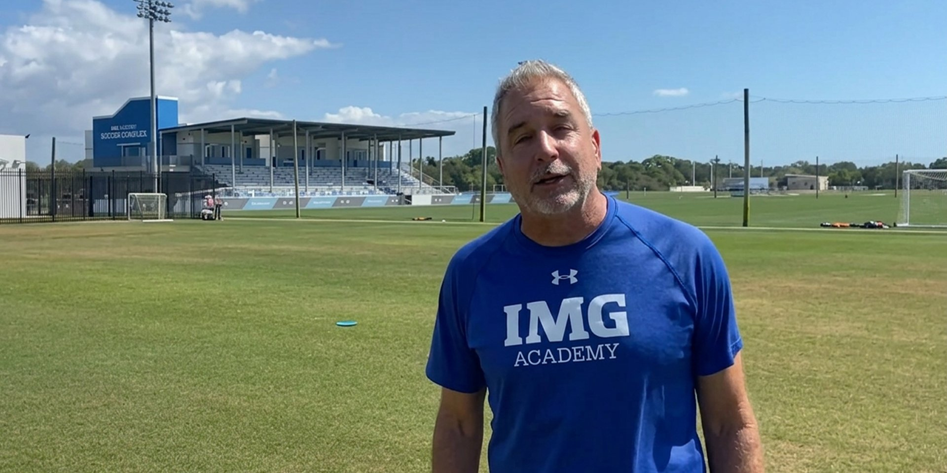 A soccer coach stands on a sunny field.