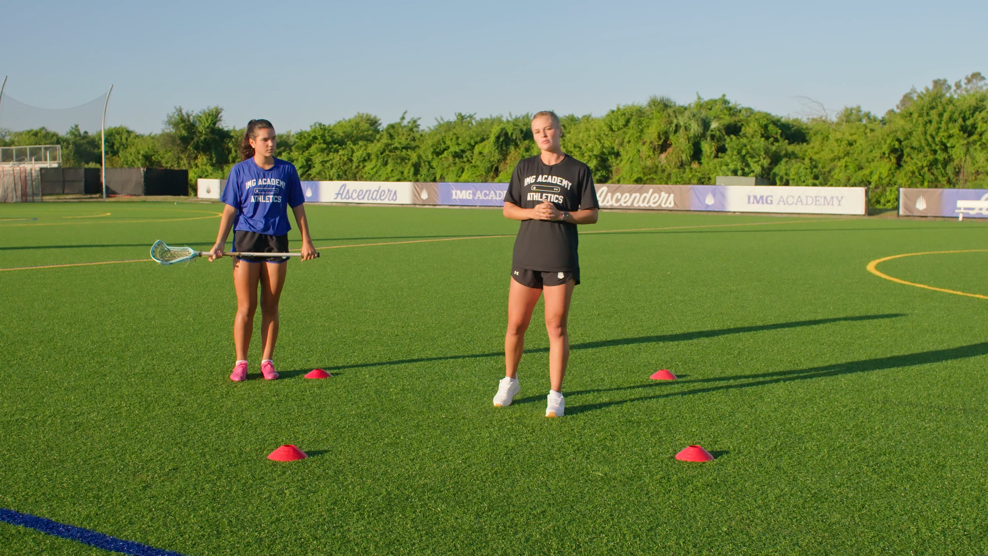 A female coach in a black athletic shirt and a female lacrosse player in a blue shirt stand on a green turf field during a sunny day. Four red cones are arranged in a square on the turf between them. The player on the left holds a lacrosse stick horizontally at her waist.