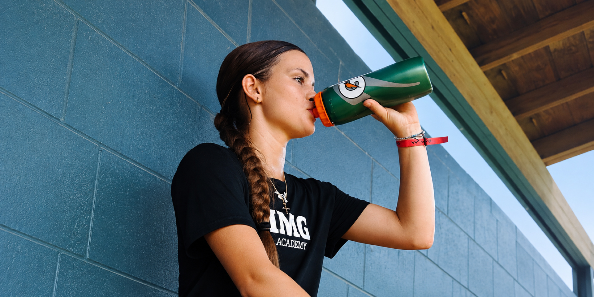 An athlete drinks from a green bottle beside an indoor court.