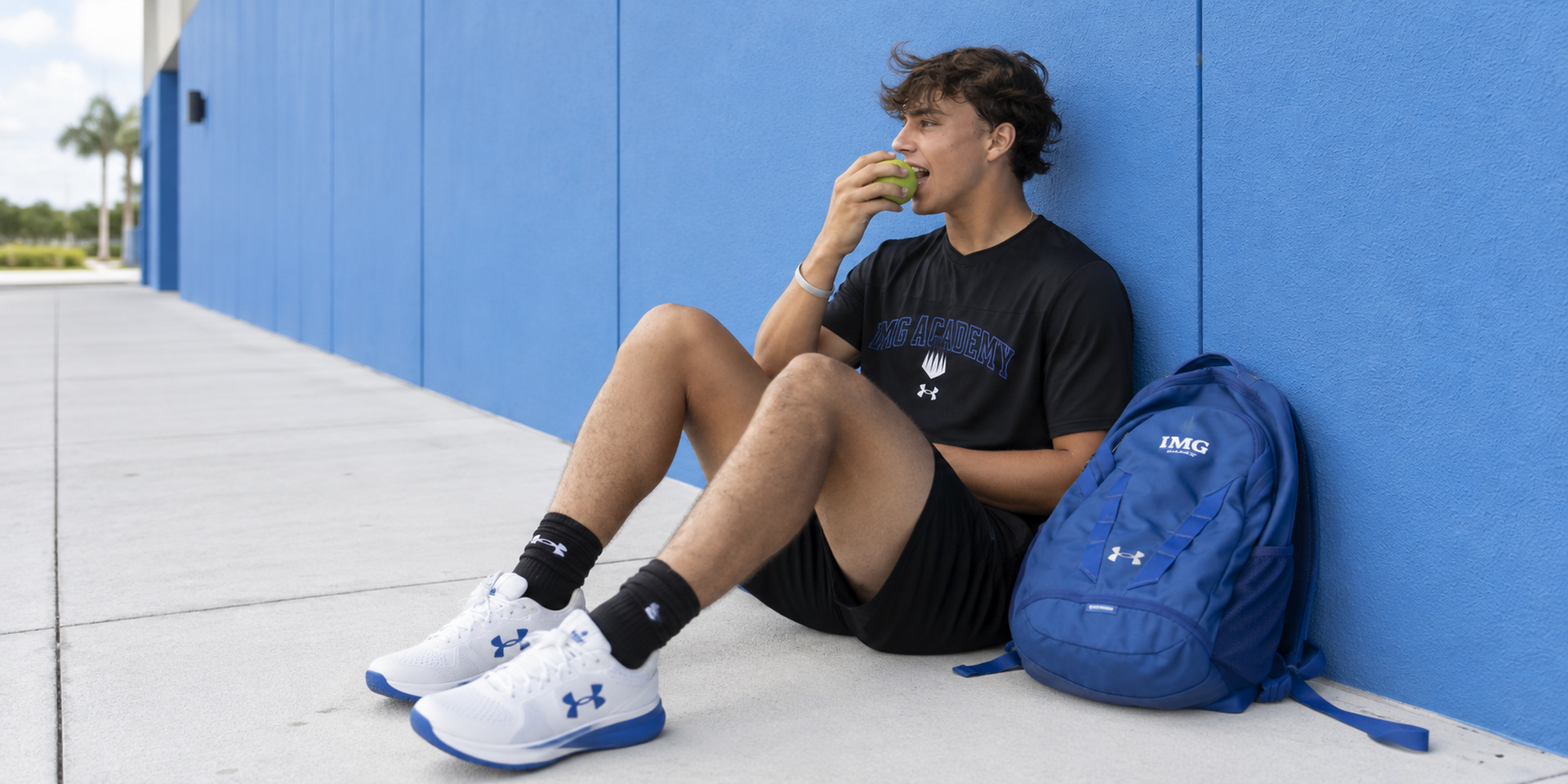 An athlete sits beside a blue wall eating a snack with a backpack nearby.