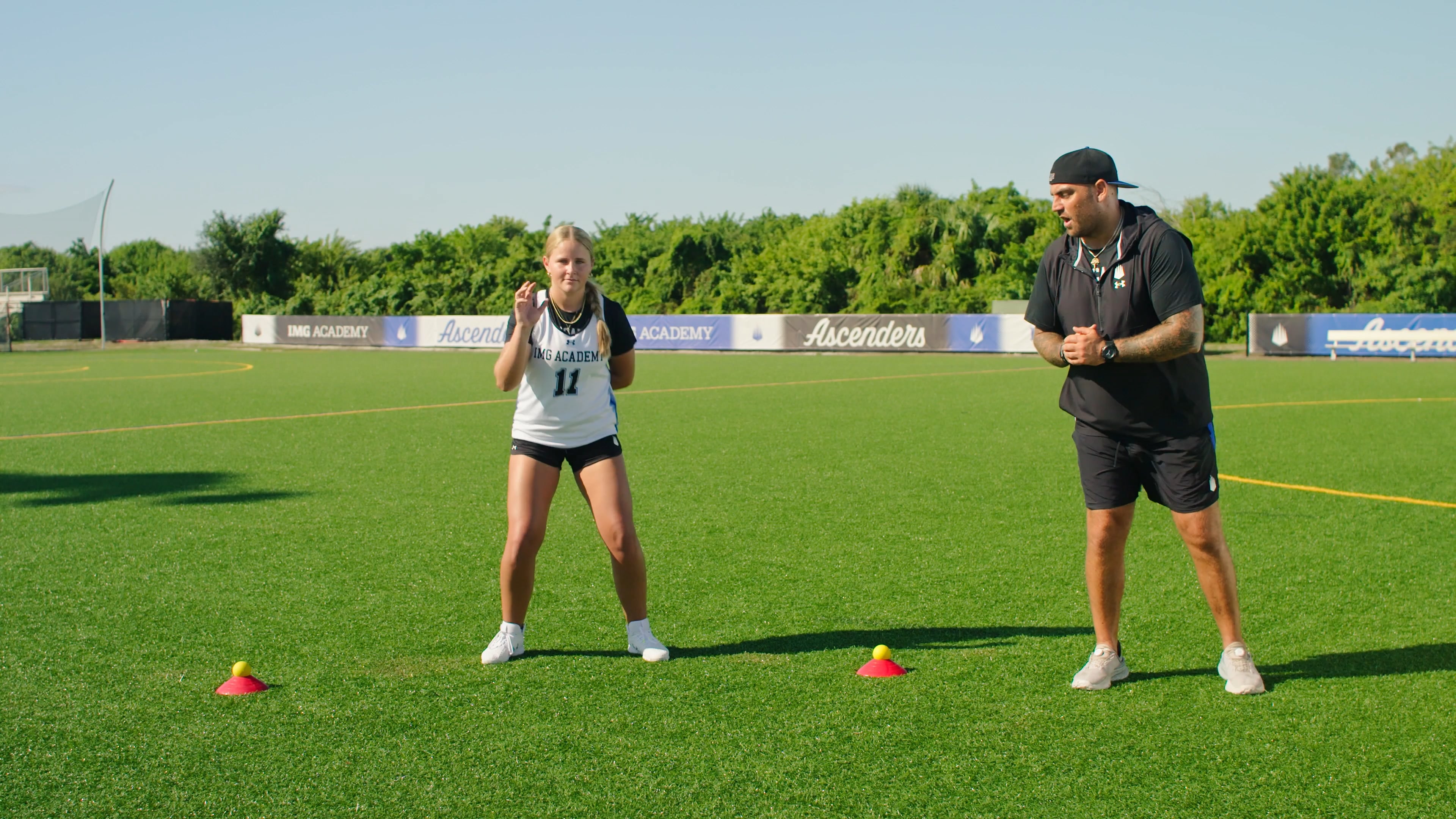 A female athlete in a white IMG Academy jersey stands in an athletic stance on a green turf field during a drill. A coach stands opposite her, providing instruction near red cones topped with yellow balls.