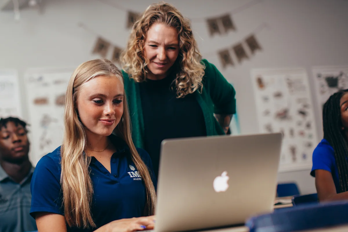 Student-athlete researching colleges on a laptop with guidance from a teacher, illustrating the support available during the college search process.