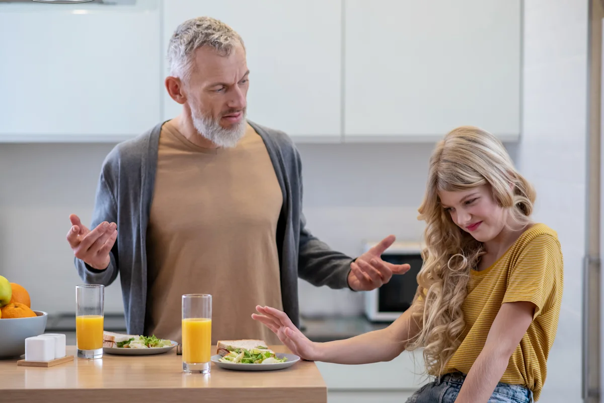 Frustrated father with his young, long-haired daughter who is pushing away a healthy meal (sandwich) in a modern kitchen, illustrating picky eating challenge.