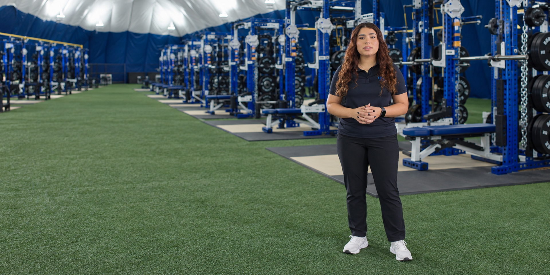 A trainer stands on indoor turf beside weight-training equipment.
