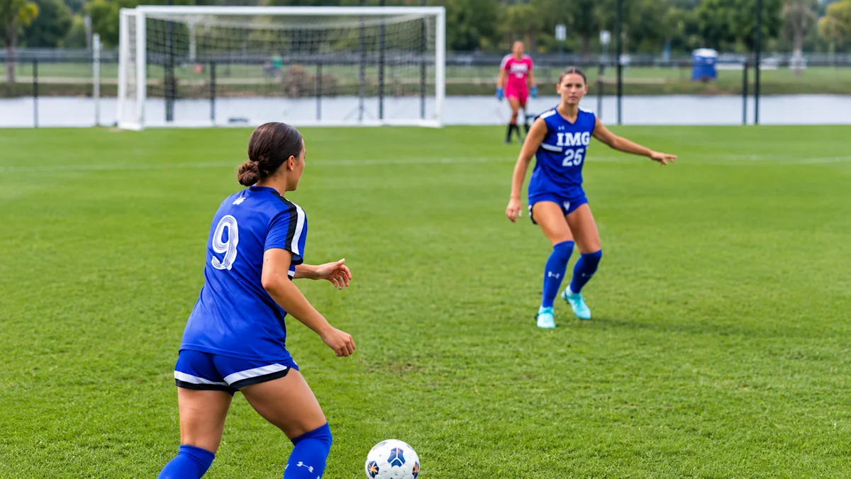 IMG Academy female soccer players in blue jerseys on a grass field, focused on a game play.