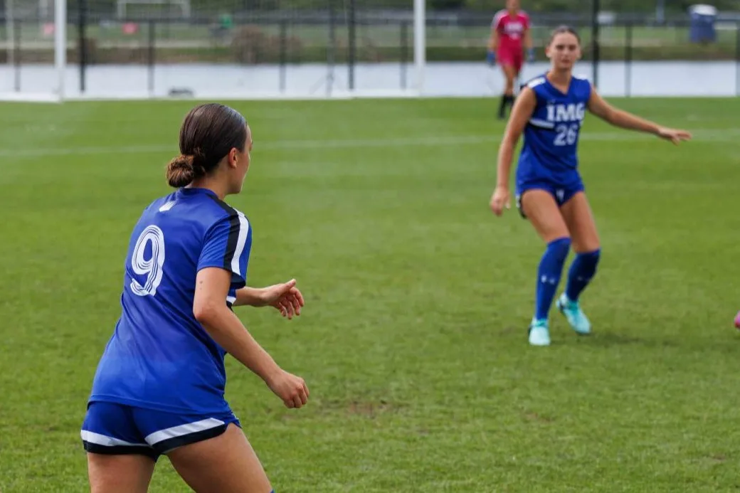 IMG Academy female soccer players in blue jerseys on a grass field, focused on a game play.