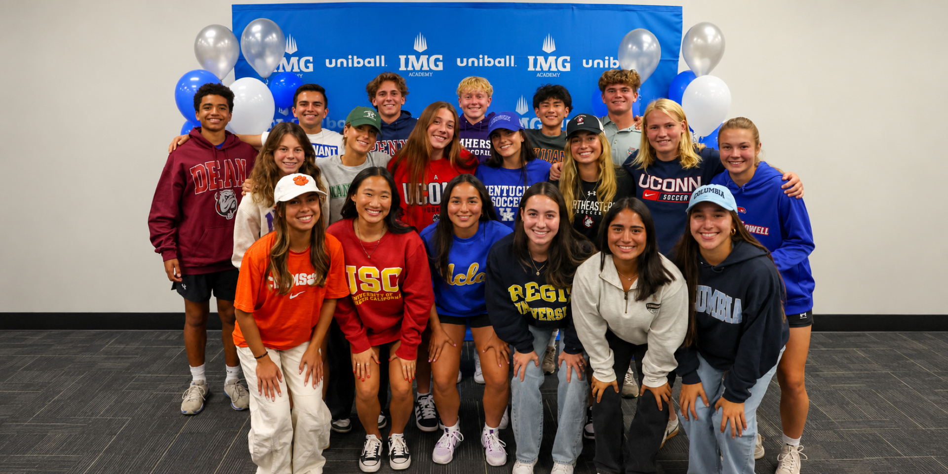 A group of student-athletes poses in college shirts in front of an IMG Academy backdrop.