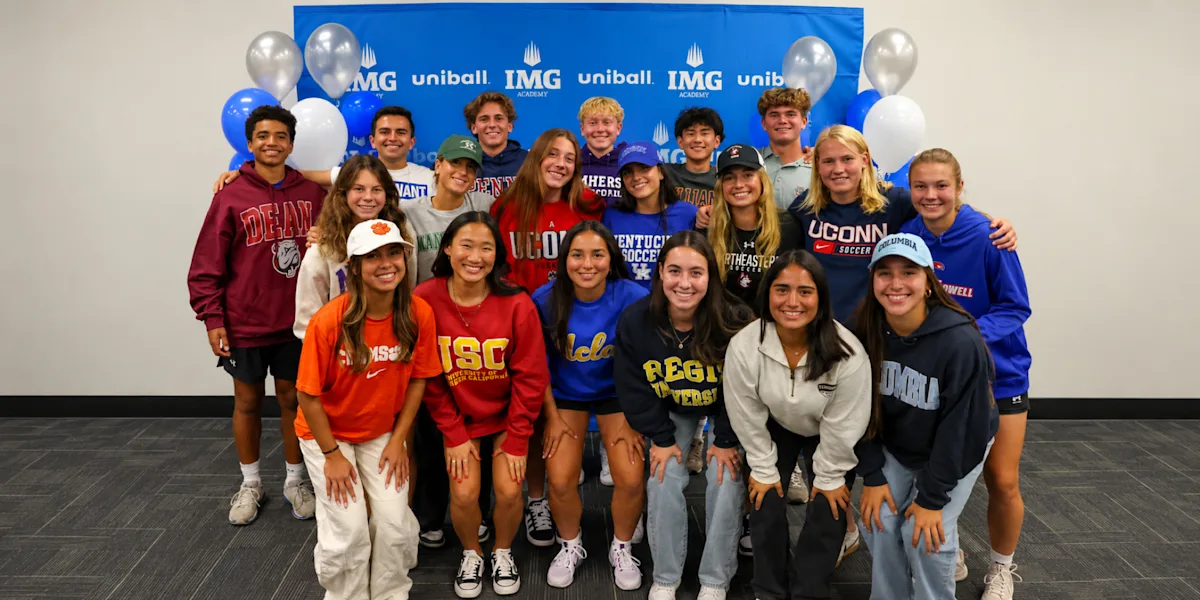 A group of student-athletes poses in college shirts in front of an IMG Academy backdrop.