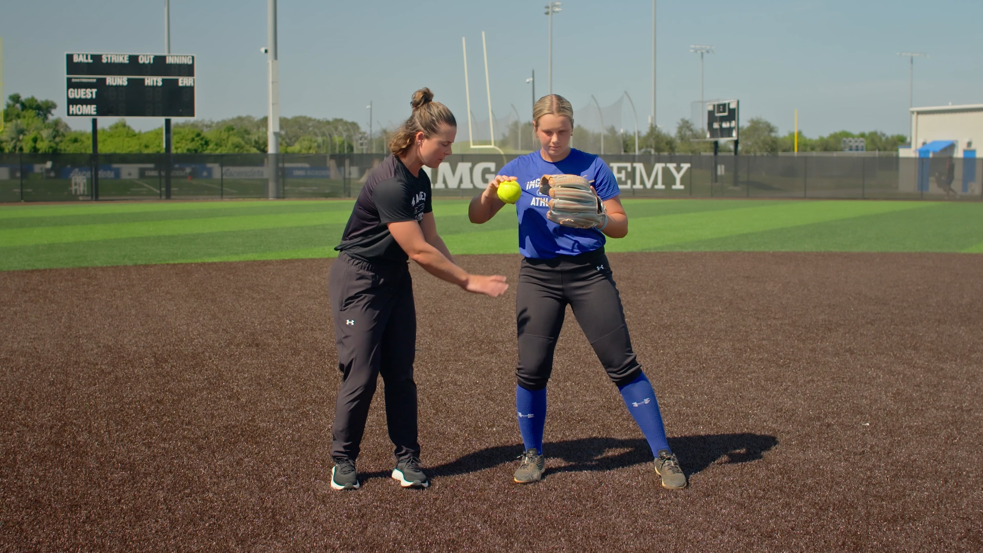 A coach in black athletic wear instructs a softball player in a blue jersey. They are standing on a dirt infield with a green outfield and a scoreboard in the background.