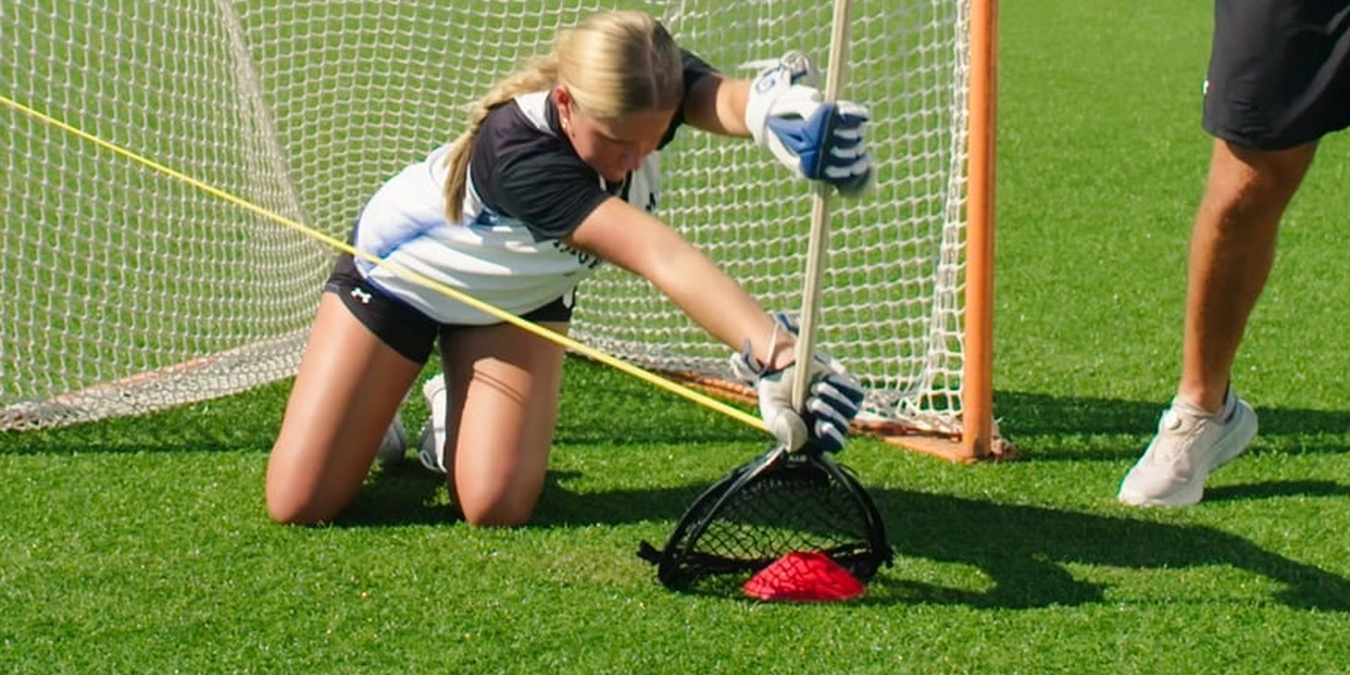 A lacrosse goalie kneels and reaches her stick across the goal mouth.