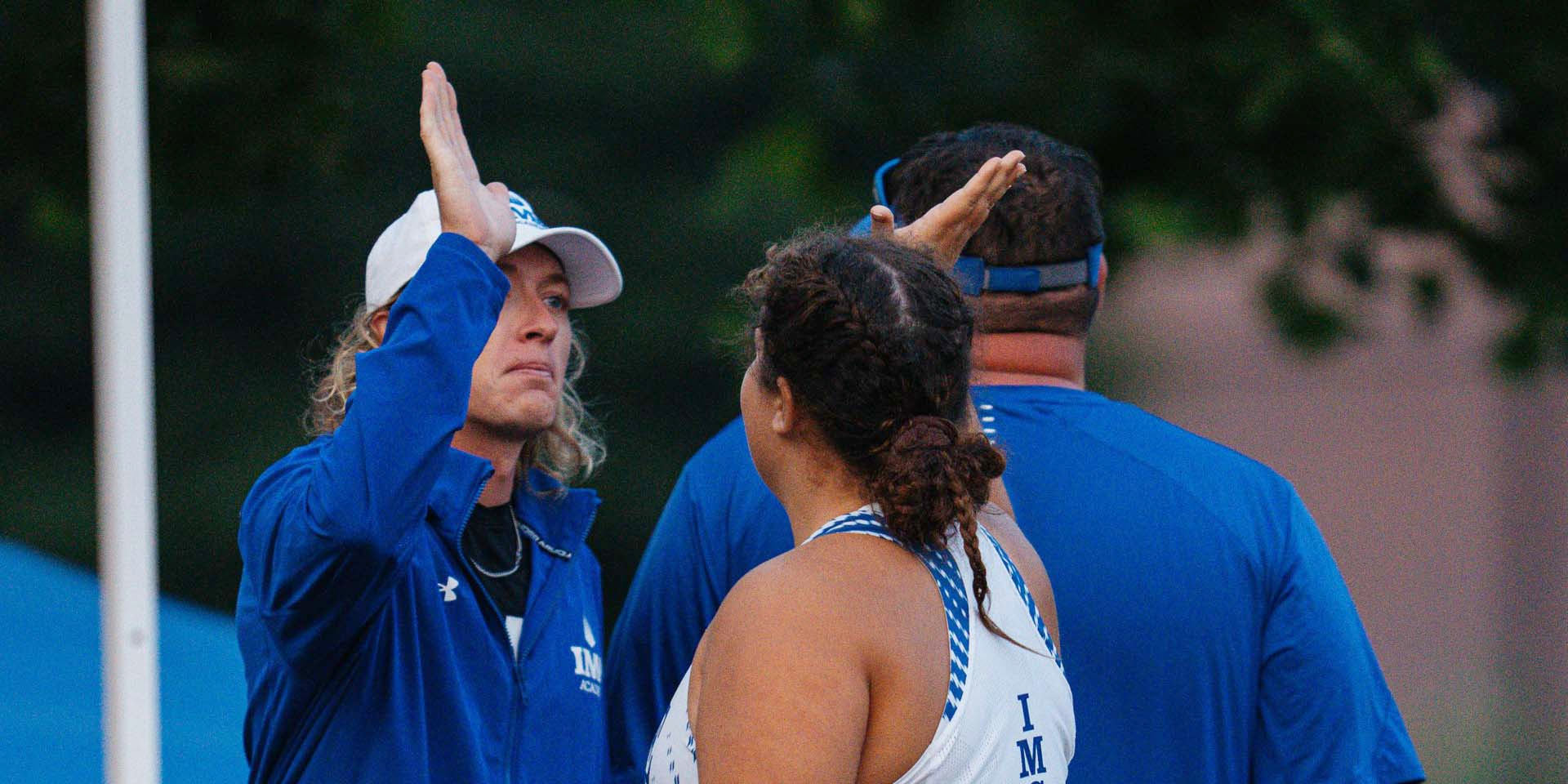 A tennis coach raises a hand while speaking with a player.