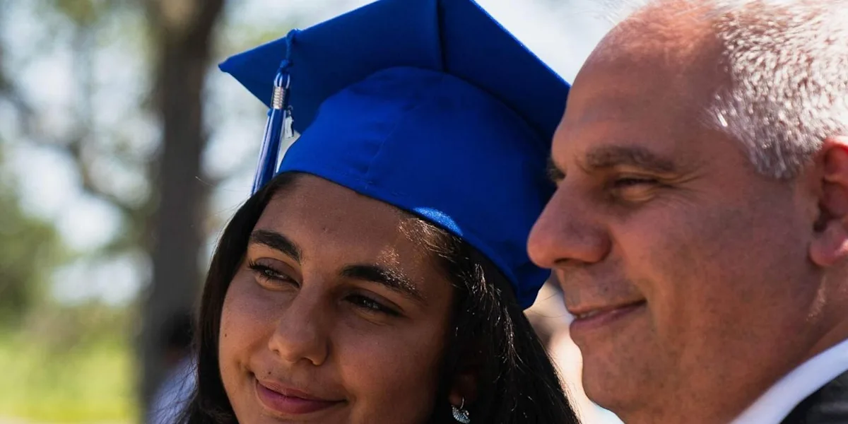 A graduate in a blue cap and gown smiles beside an adult.