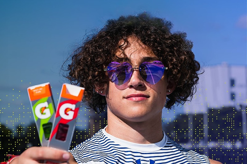 IMG Academy athlete with curly hair wearing reflective sunglasses holding two Gatorade energy chews outdoors, representing hydration, fueling, and performance nutrition.