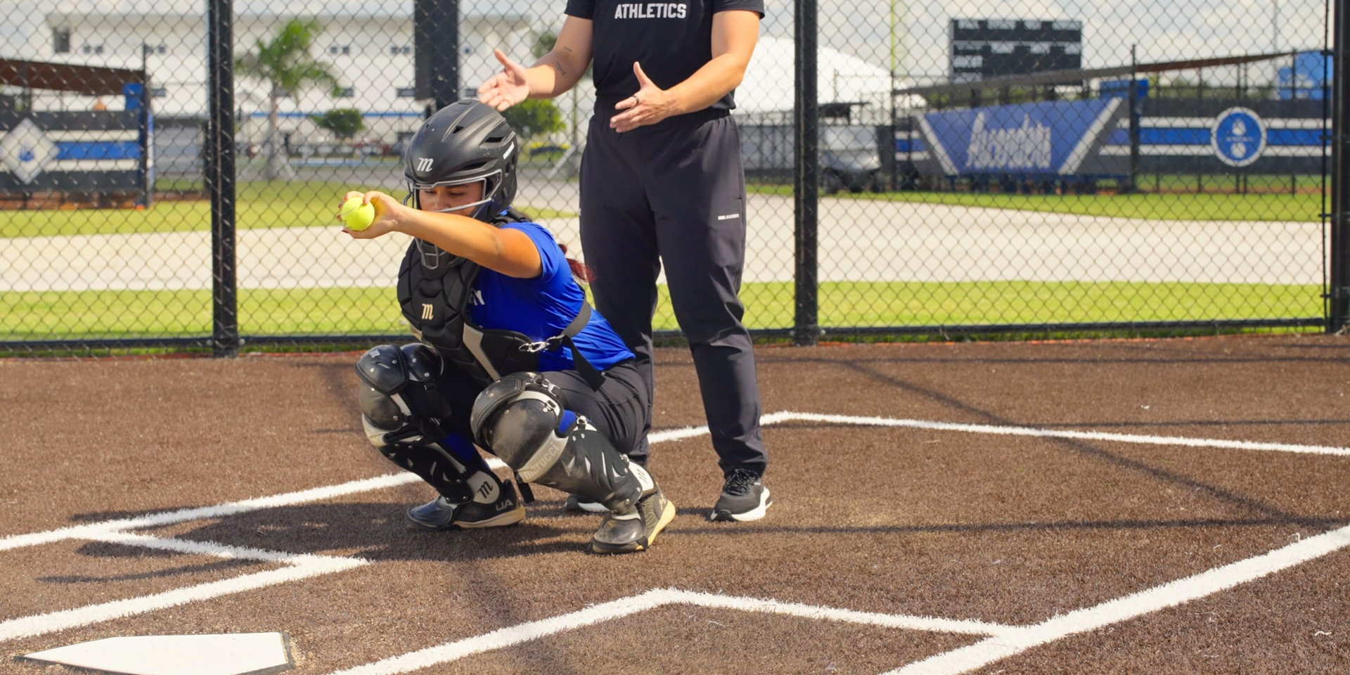 A catcher crouches at home plate holding a tennis ball while a coach observes.