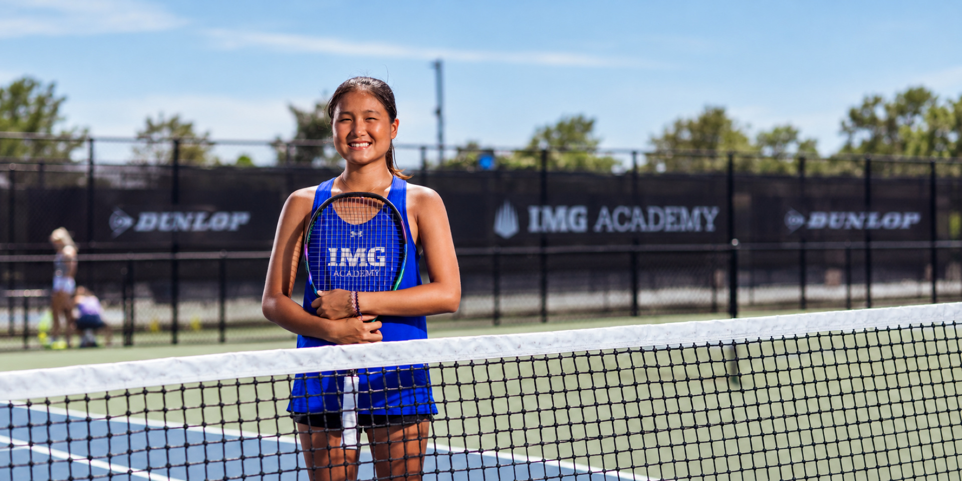 A tennis player stands smiling behind the net on an outdoor court.