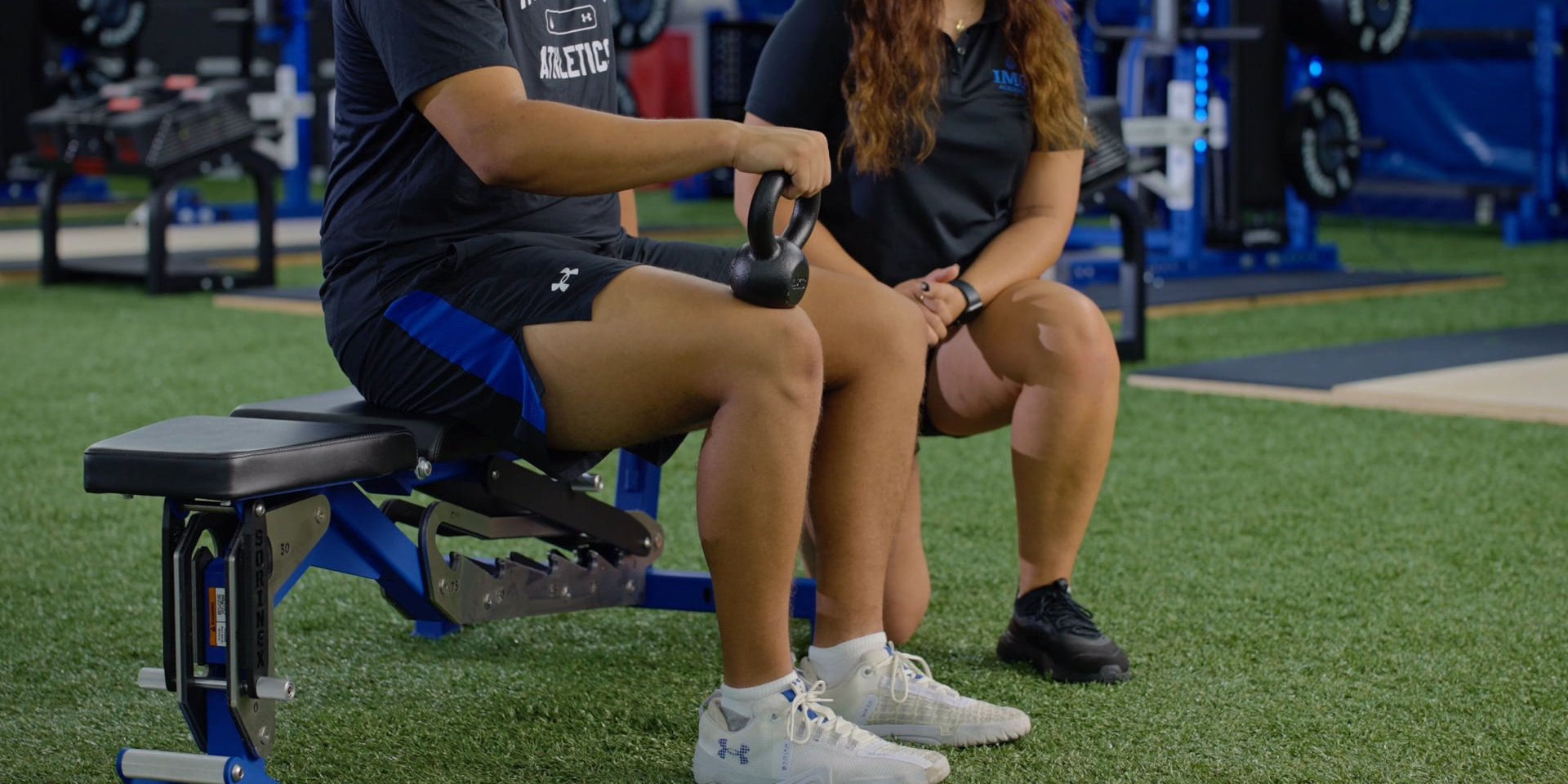 A seated athlete lifts one knee while holding a kettlebell beside a trainer.