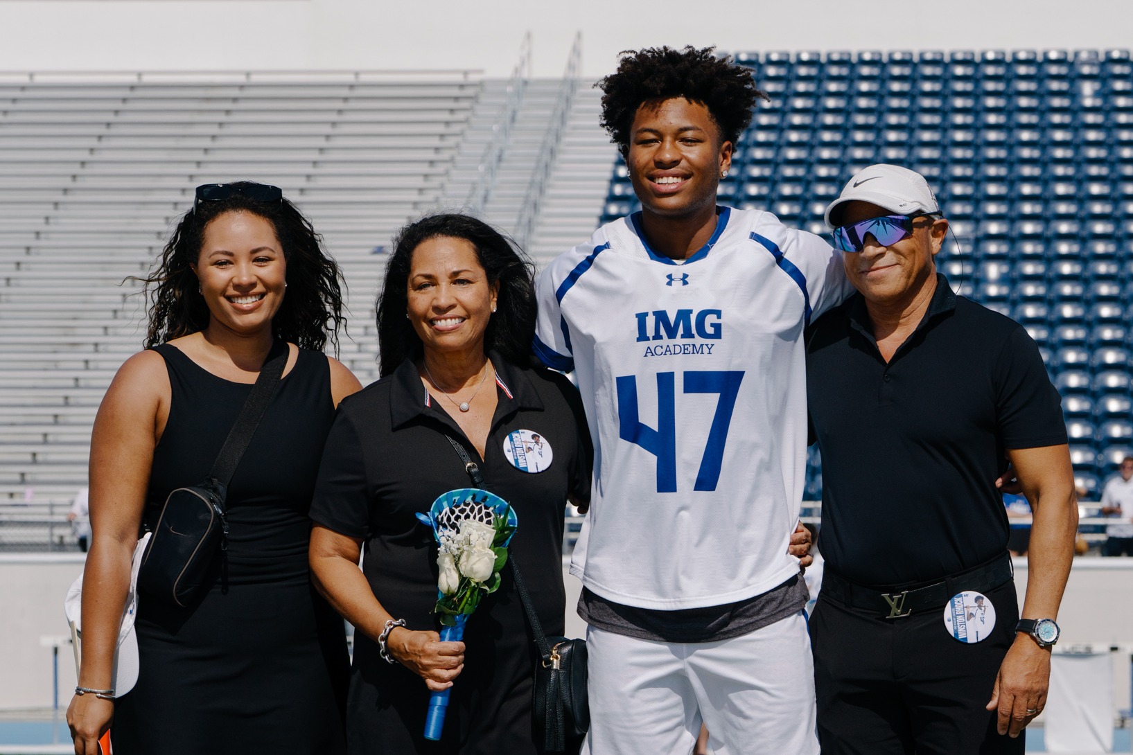 Lacrosse athlete (jersey #47) smiling with his family (parents and sister) on the turf during Senior Night ceremonies at IMG Academy Field.
