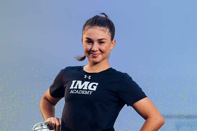 IMG Academy female student-athlete standing confidently with hands on hips, smiling while wearing an IMG Academy shirt against a clear blue sky.