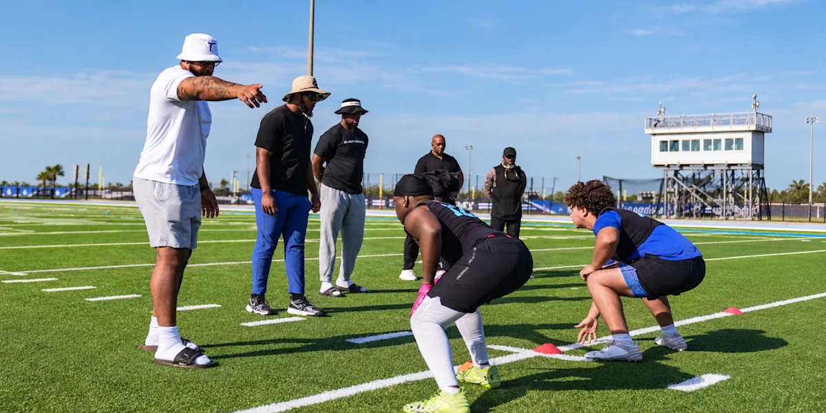 Coaches observe football players practicing on a field.