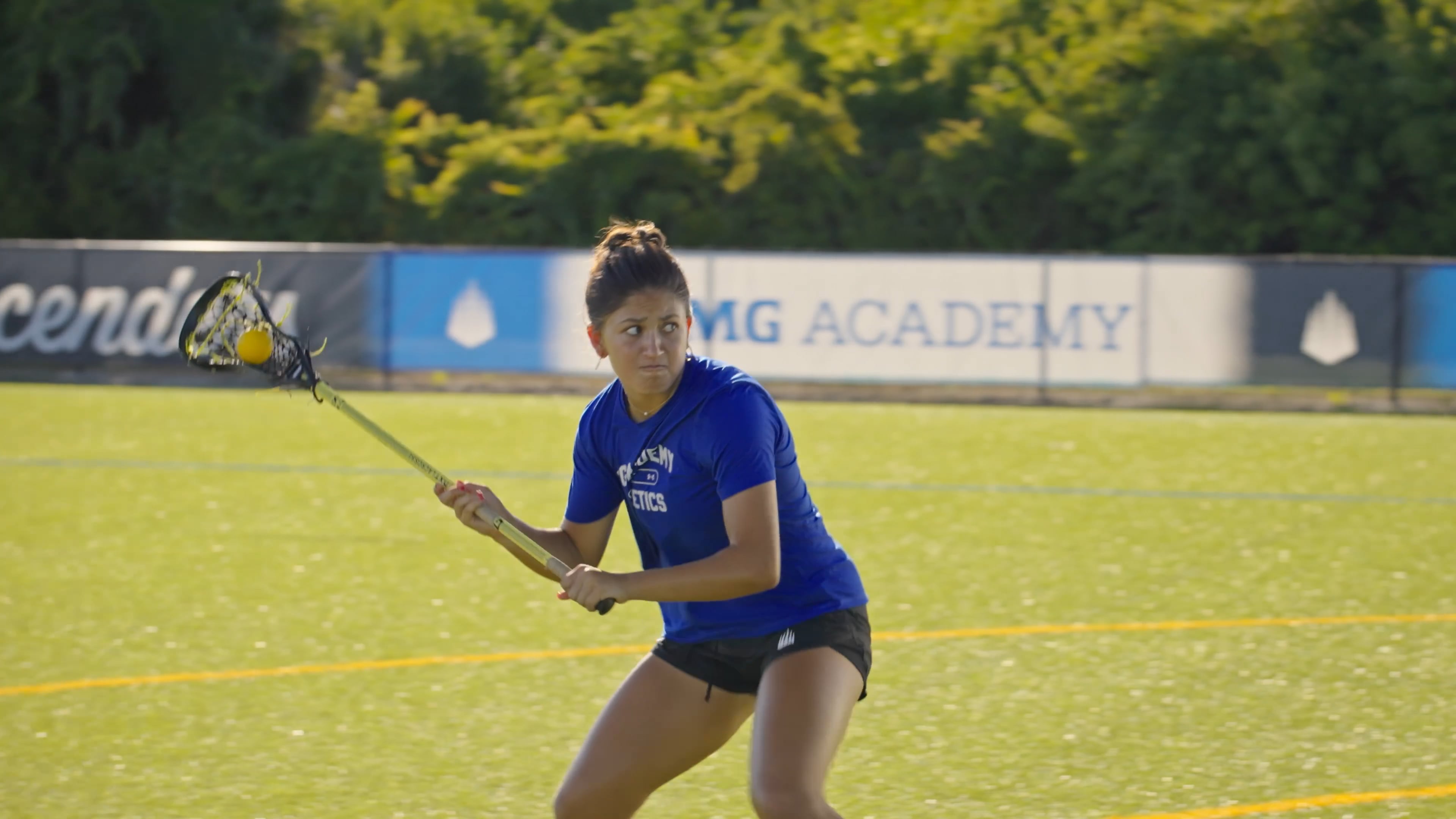 A female lacrosse player in a blue athletic shirt and black shorts moves across a green turf field, holding her lacrosse stick with a yellow ball in the net. She is in a focused, crouched athletic stance.