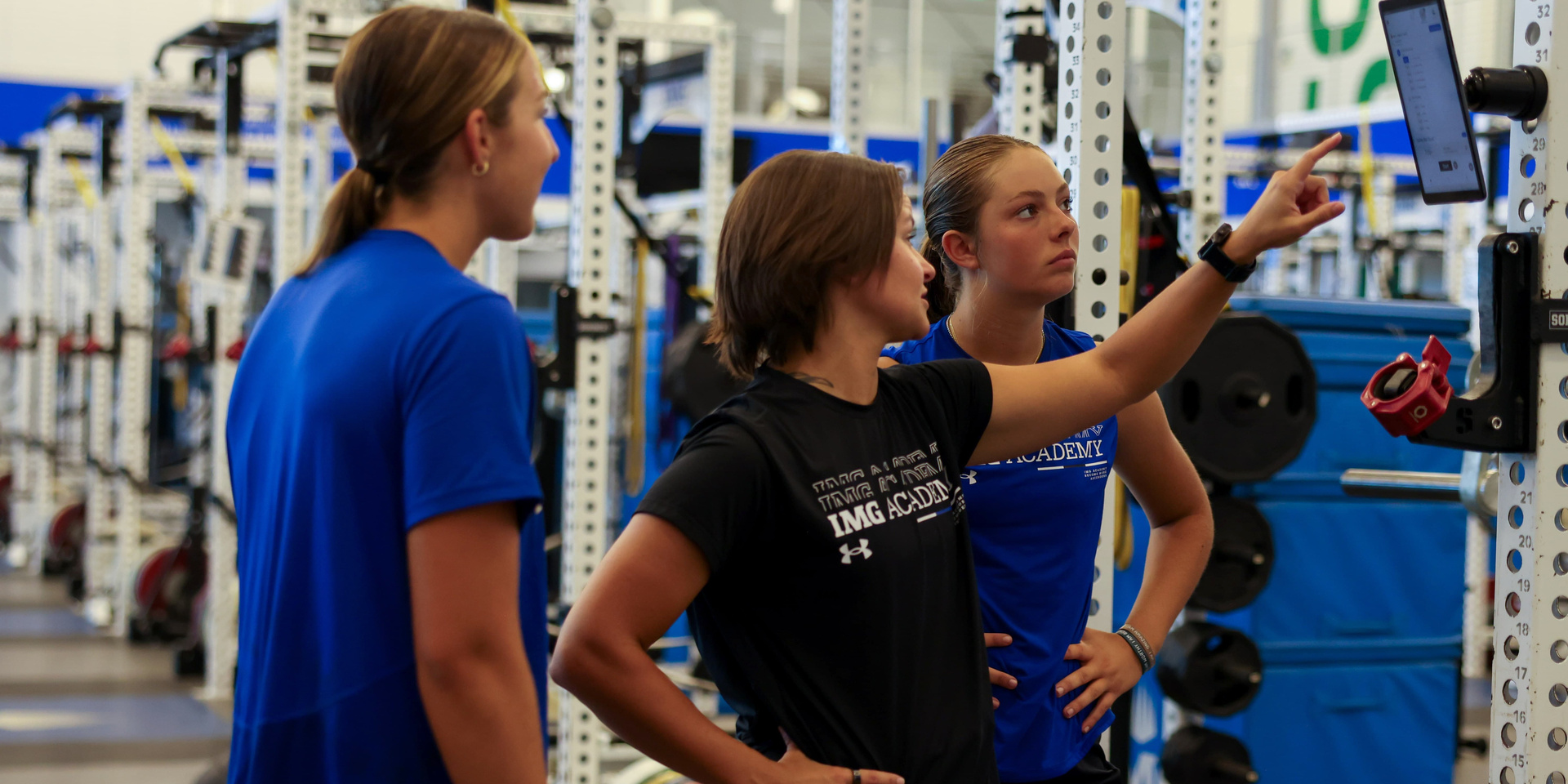Three student-athletes look at a tablet mounted on a weight-room rack as one points at the screen.