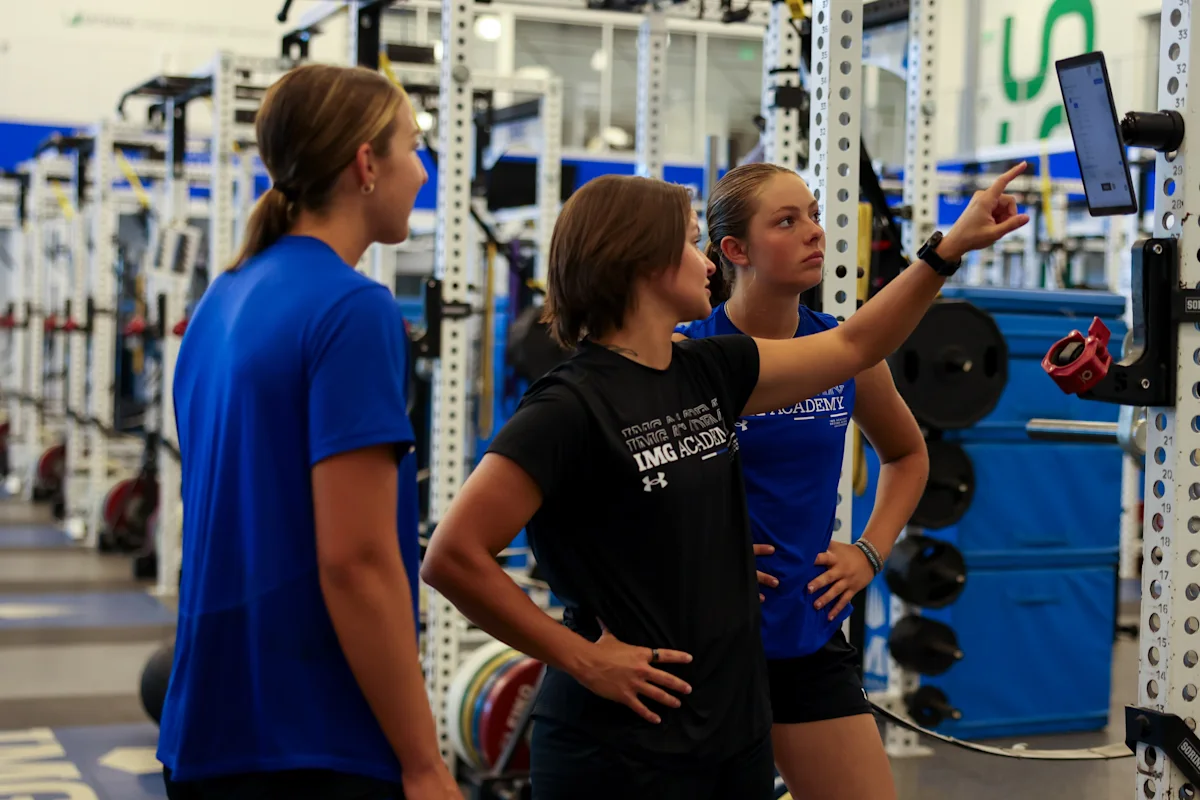 Three IMG Academy student-athletes stand in the weight room, looking at a digital screen mounted on a training rack to review strength metrics, workout plans, and overall goals.