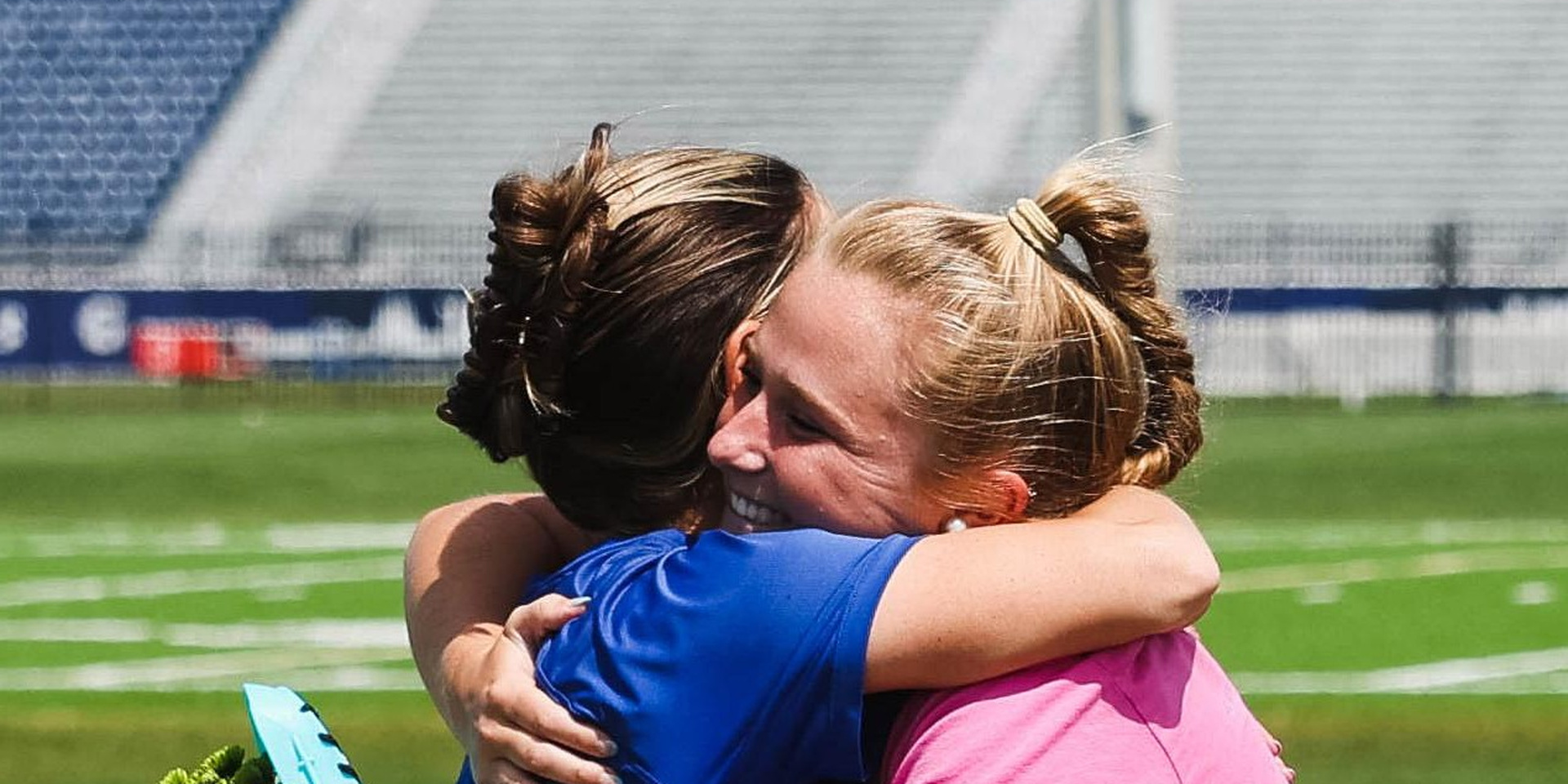 An athlete and an adult embrace beside a sports field.