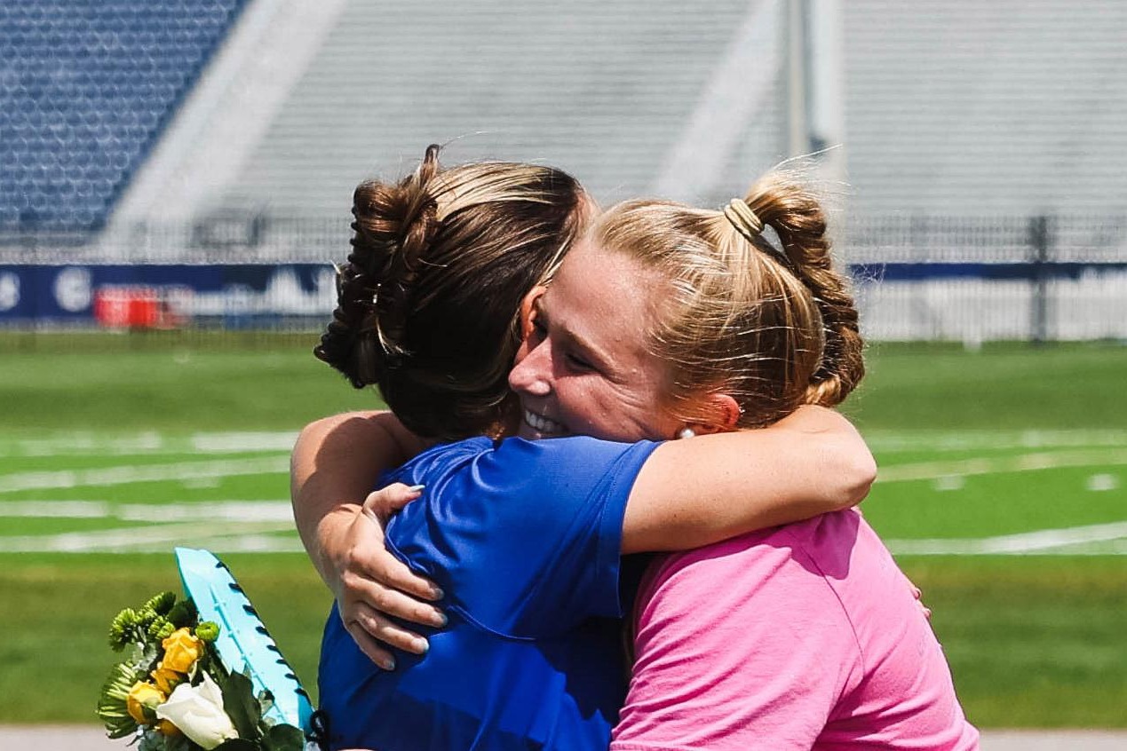 A mom and female athlete sharing an emotional hug on a sports field during a graduation or senior night ceremony.