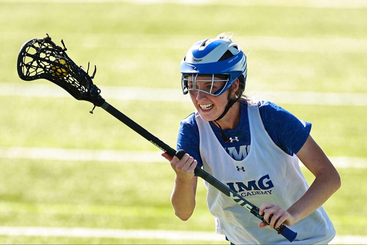 Female lacrosse player from IMG Academy cradling the ball during a game on a sunny field.