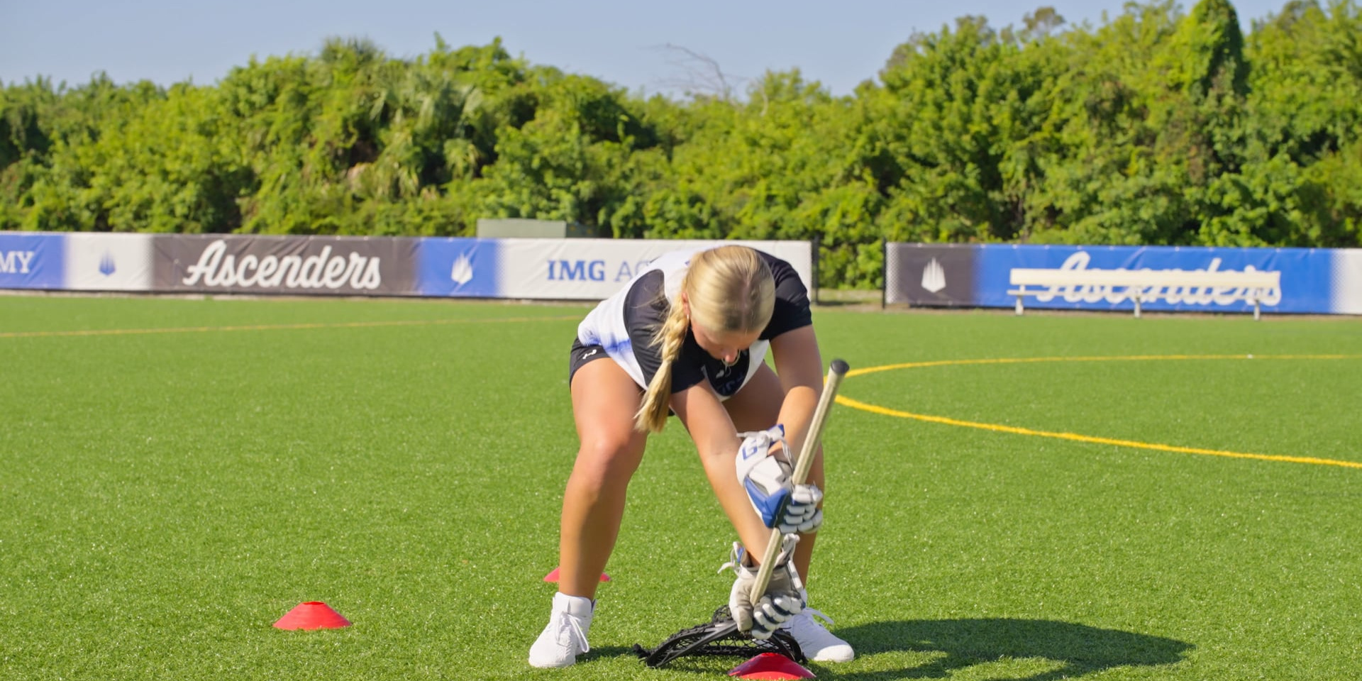A lacrosse goalie bends forward to scoop a ball with her stick.
