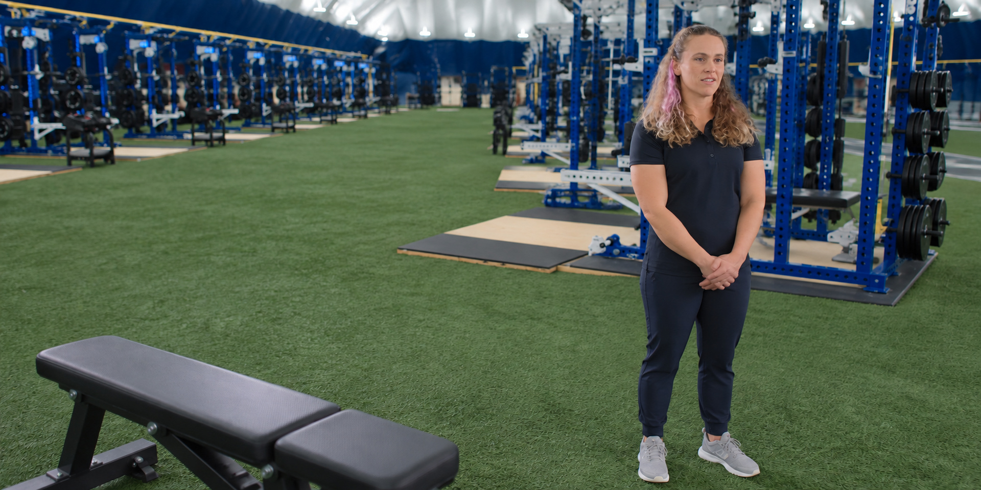A trainer stands on indoor turf beside a bench and weight racks.
