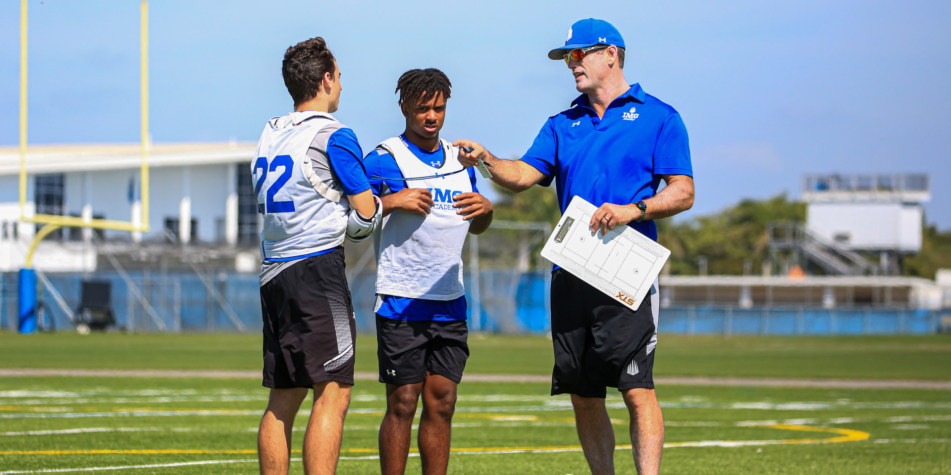 A coach holding notes speaks with two football players on a field.