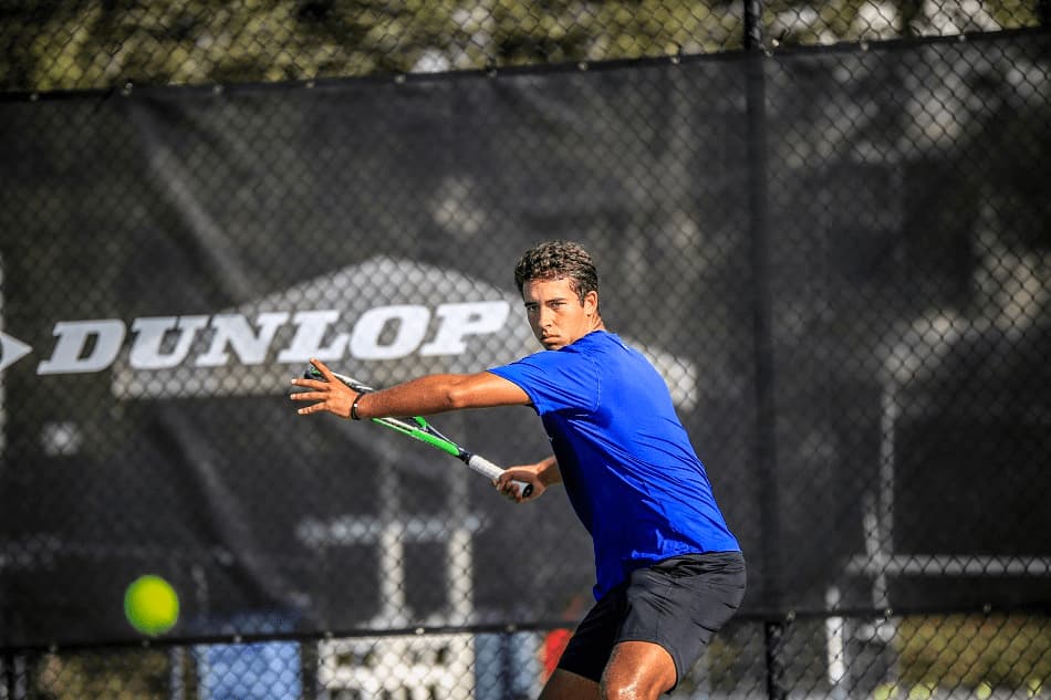 A male tennis player prepares for a forehand shot on an outdoor court, eyes focused on the incoming ball, with a Dunlop banner in the background.