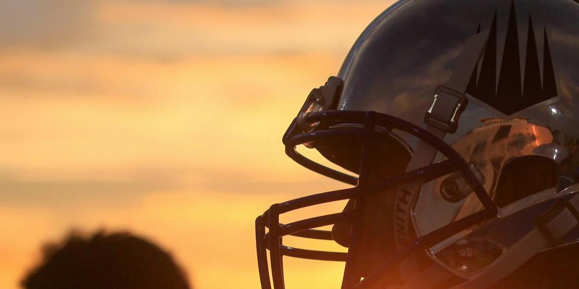 A close view shows a football helmet silhouetted against an orange sunset.