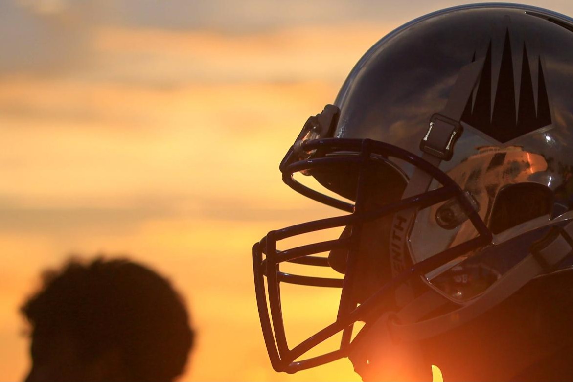 Football player in reflective helmet with black logo silhouetted against a sunset sky.