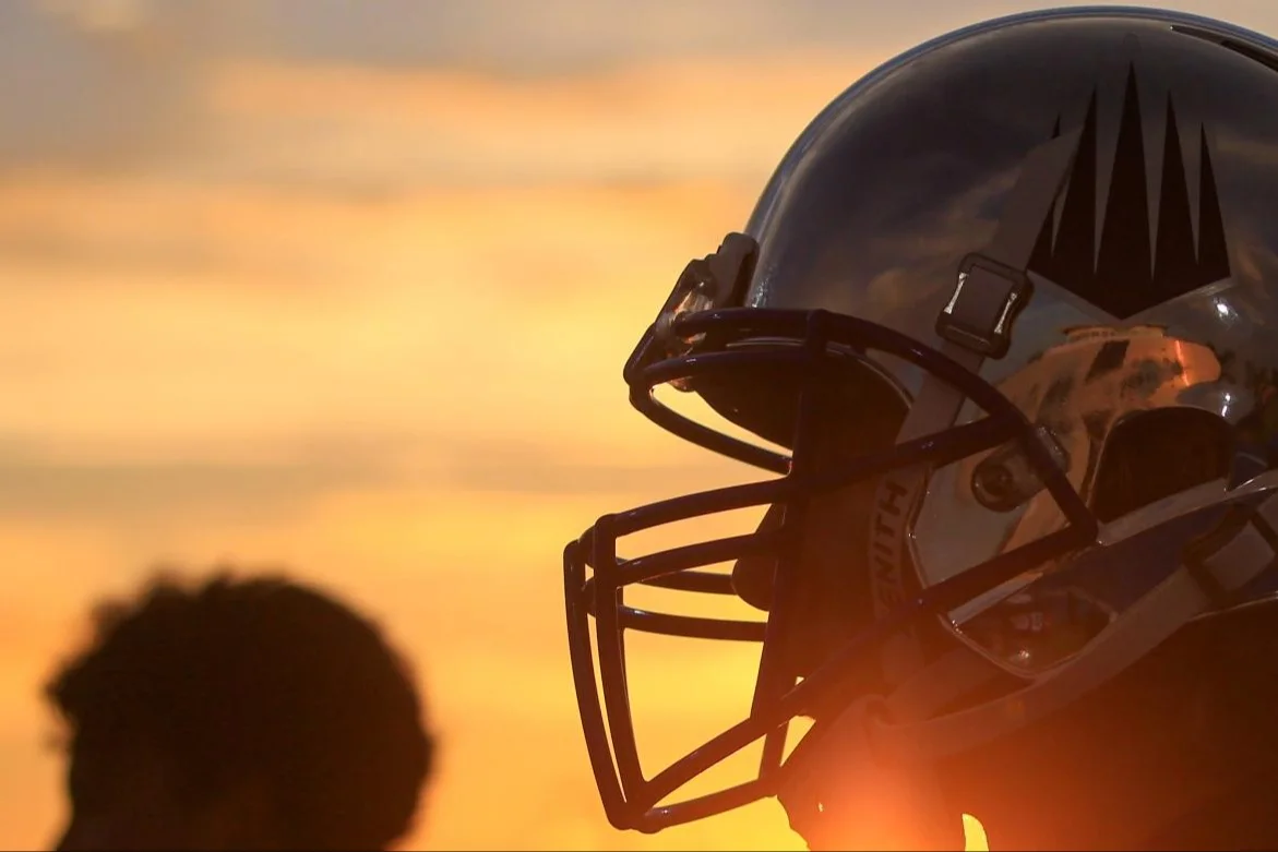 Football player in reflective helmet with black logo silhouetted against a sunset sky.