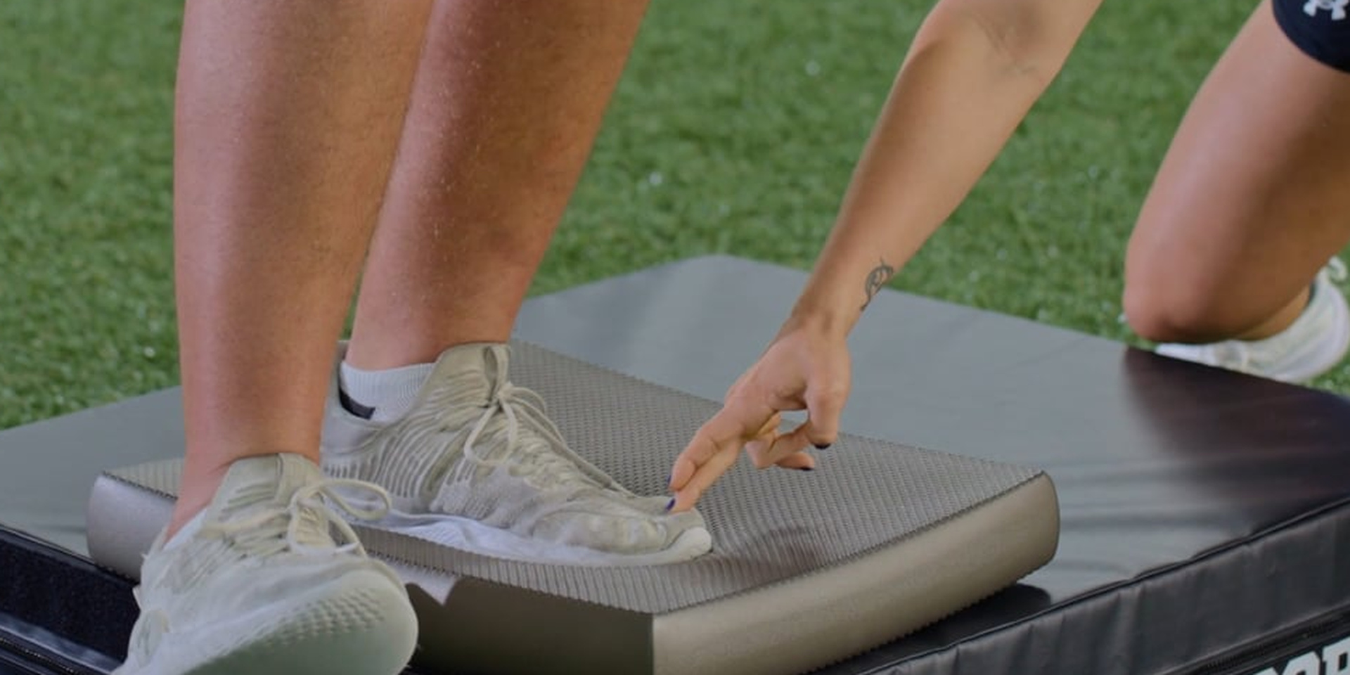 A trainer points to an athlete’s foot during a balance exercise on a padded step.