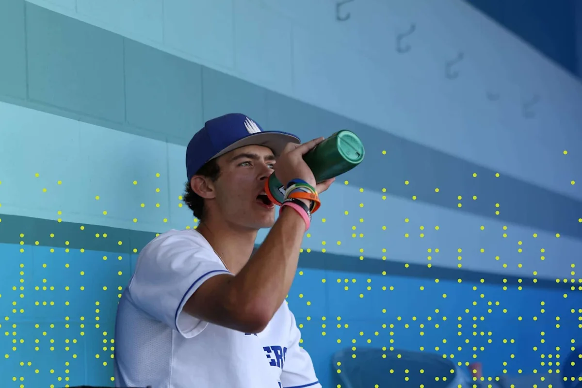 Baseball player in a dugout drinking from a water bottle, wearing a cap and wristbands.