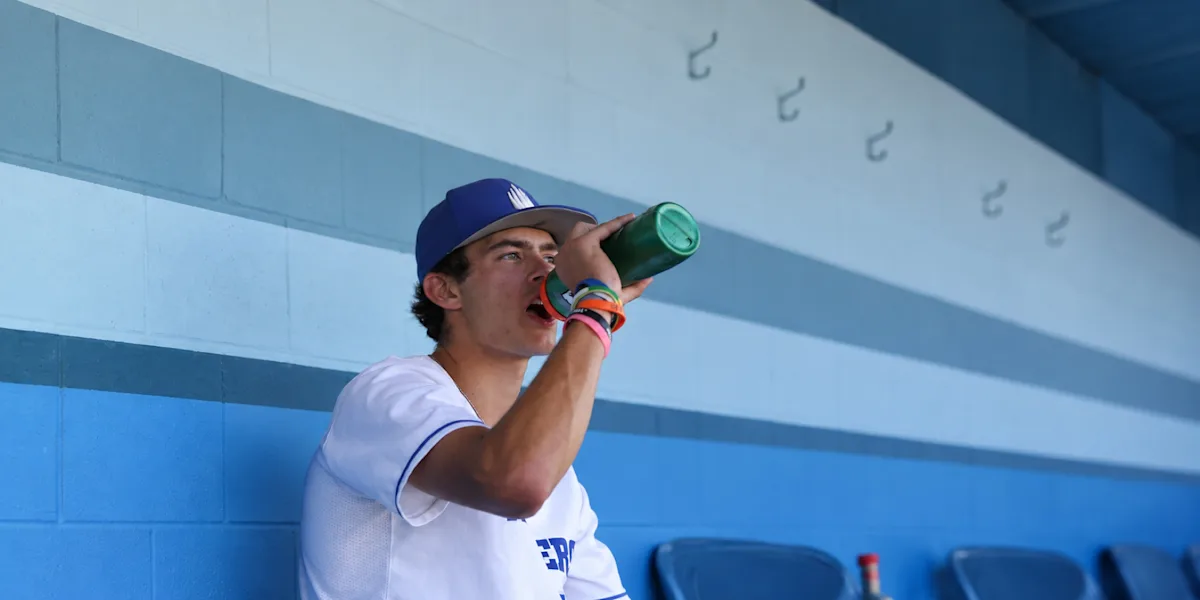 A baseball player drinks from a green bottle in a dugout.