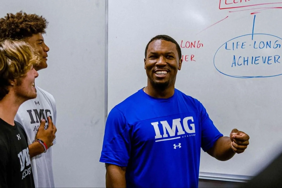 Coach leading a group of student-athletes in a classroom at IMG Academy, standing in front of a whiteboard with the phrase "Life-Long Achiever" written on it.