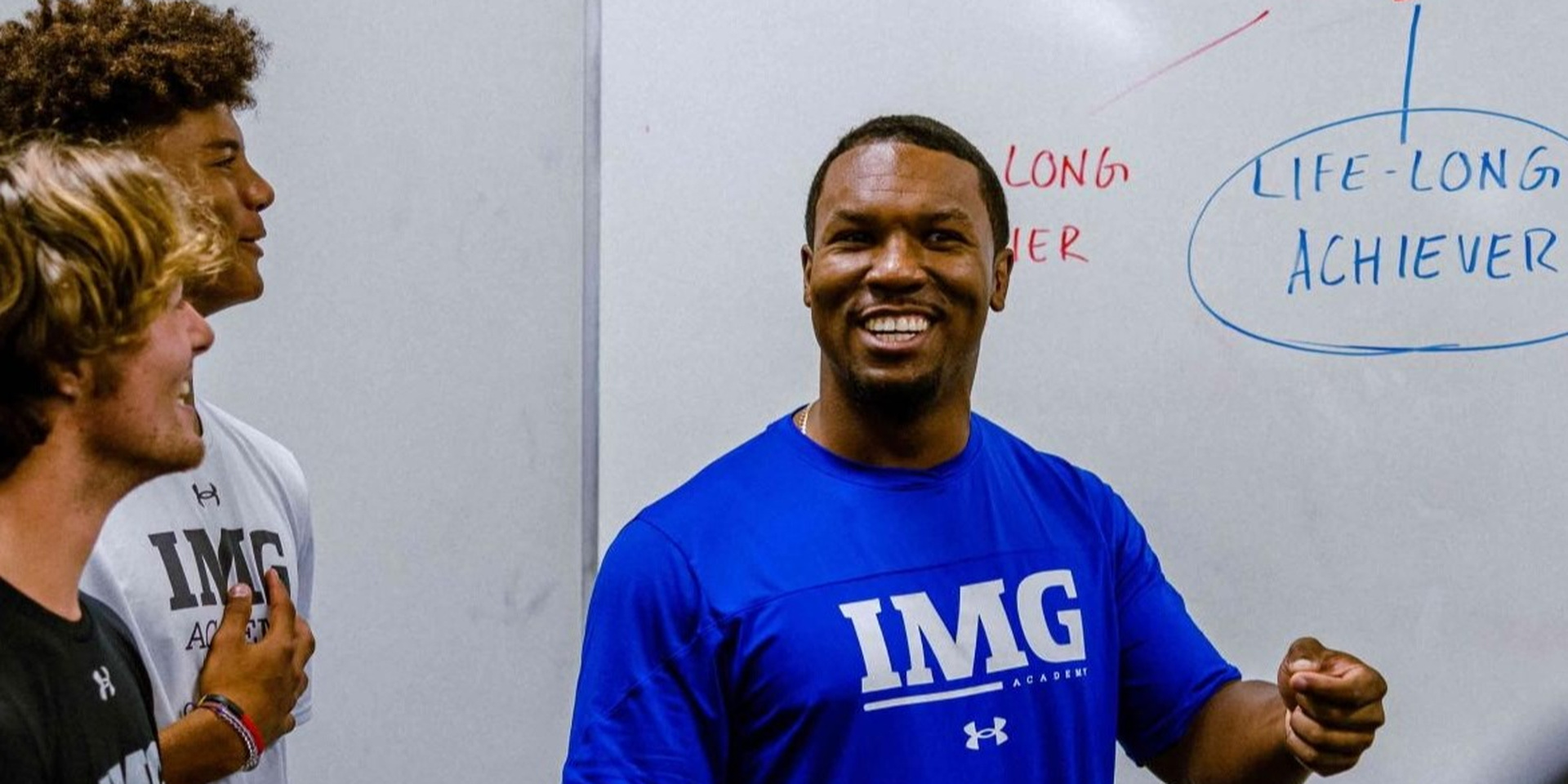 A smiling coach speaks beside a whiteboard in a classroom.