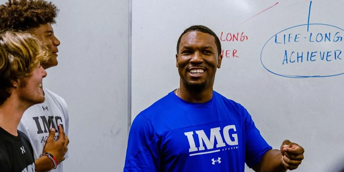 A smiling coach speaks beside a whiteboard in a classroom.