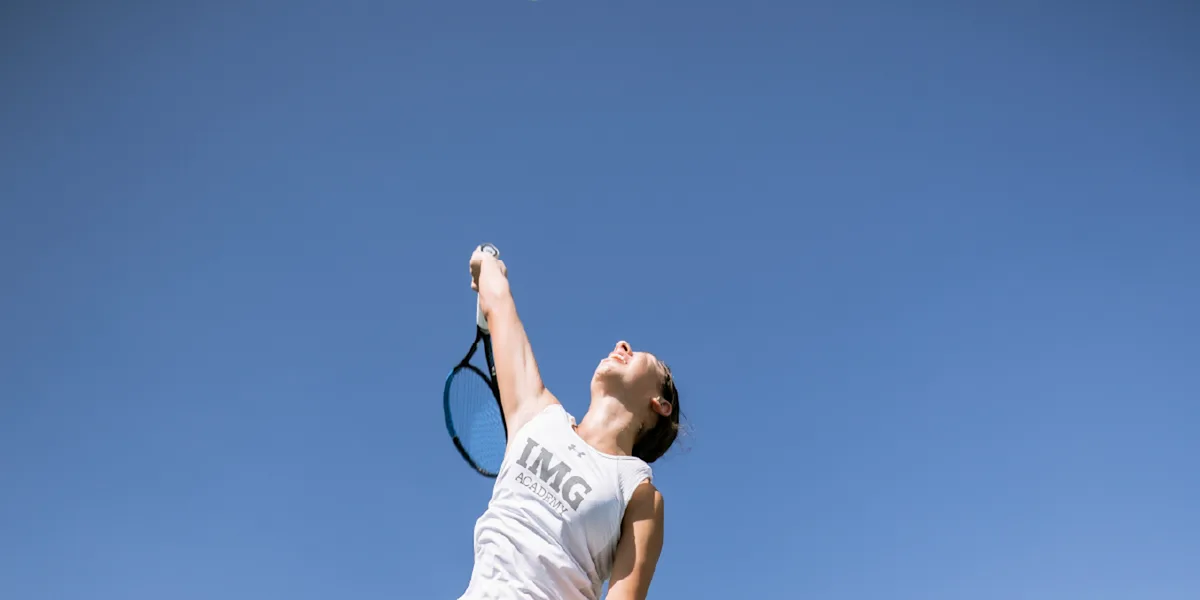 A tennis player stretches upward to serve against a blue sky.