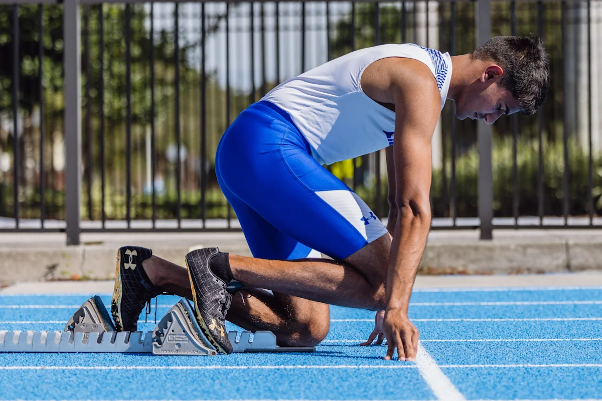 Male IMG Academy track athlete kneels at the starting line on a bright blue track, preparing for a sprint and focusing on the start of the race or practice.