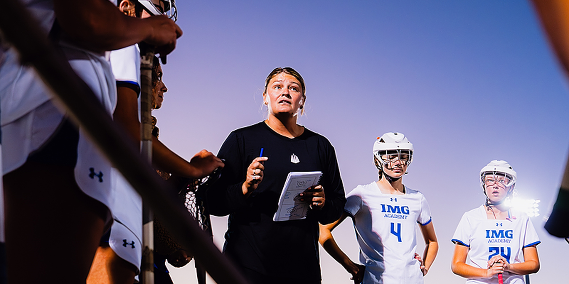 A coach holding a clipboard addresses a group of lacrosse players on an outdoor field.