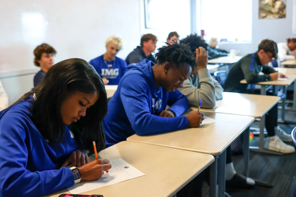 IMG Academy students sit at desks in a bright classroom, focused on writing during a sports marketing or academic exercise, wearing branded blue athletic apparel.