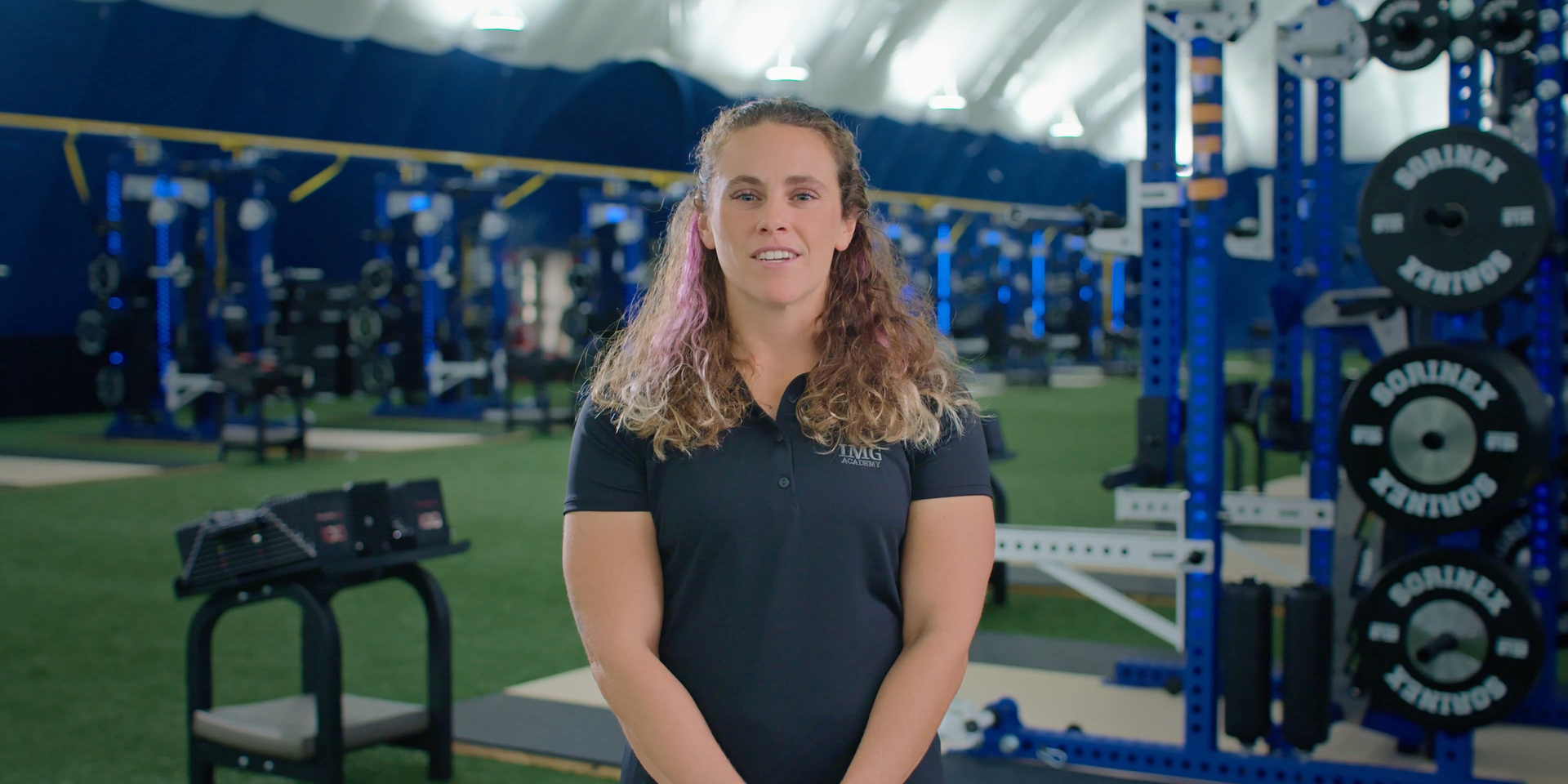 A trainer stands on indoor turf surrounded by strength-training racks and equipment.
